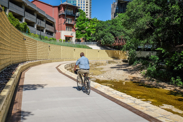 Cyclist riding on a concrete hike and bike path with a large retaining wall and high-rise buildings on the left.