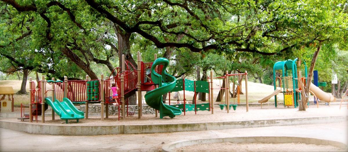 Large playscape among big oak trees with green twisty slide in the middle and a swing set.