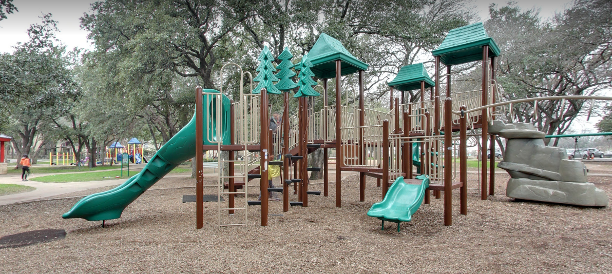 Green playscape with oak trees surrounding it, another yellow playscape is in the background.