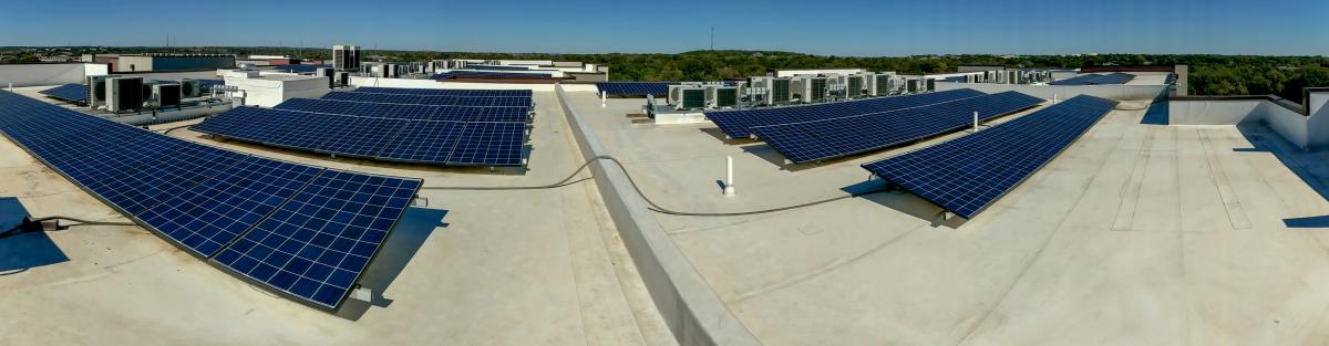Panoramic view of a large rooftop solar array under a blue sky. Trees and hills are on the horizon line.